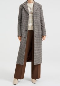 Lång rutig kappa i svart och vitt, med stora slag, knäppning och en avslappnad passform, över en krämfärgad stickad tröja och bruna byxor.