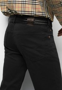 Persoon in zwarte jeans met achterzakken, gevlochten zwarte riem met een leren label "M5" en een beige geblokt overhemd dat is ingestopt.