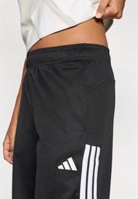 Črne športne hlače iz teksturirane tkanine z elastičnim pasom, okrašene z belimi tremi črtami in logotipom Adidas.