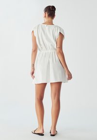 Weißes kurzes Kleid mit tailliertem Bund, kurzen Ärmeln und einem rückseitigen Schlüssel-Ausschnitt. Glatter Stoff und lockere Passform. Schwarze Sandalen getragen.