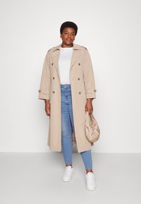 Trench coat bej cu design cu două rânduri de nasturi, mâneci suflate și nasturi mari. Purtați-o cu o cămașă albă, blugi bleumarin și adidași albi.