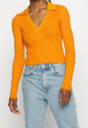 Osoba ubrana w dopasowany, pomarańczowy, prążkowany sweter z długim rękawem i dekoltem w serek z kołnierzykiem oraz jasnoniebieskie, wysokowalne dżinsy.