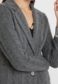 Šedý herringbone blejzr s vyvýšeným límcem, dvěma knoflíky a žebrovanými detaily na rukávech. Disponuje texturovanou tkaninou a splývavým střihem.