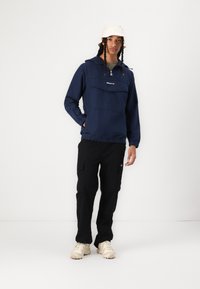 Marineblå genser med frontlomme, elastisk hem og hette, kombinert med svarte cargo-bukser og lyse beige sko.