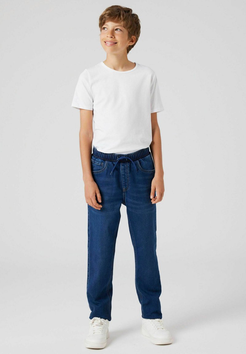 Tricou alb din bumbac combinat cu pantaloni din denim albastru, prevăzuți cu talie elastică și șnur. Model purtând adidași albi.