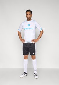 Camiseta de fútbol blanca con detalles en azul y logotipo del patrocinador, combinada con pantalones cortos negros y calcetas blancas. Modelo lleva zapatillas negras con tacos.