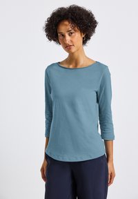 Top leggero a maniche lunghe di colore blu teal, con scollo rotondo e maniche a tre quarti, caratterizzato da una texture liscia e dettagli di cucitura discreti.