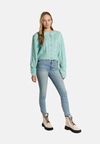 Maglione cropped in maglia a treccia verde menta con bottoni, abbinato a jeans skinny blu chiaro e stivali da combattimento beige con lacci.