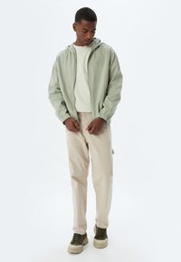 Lys grøn letvægtsjakke med hætte, elastiske manchetter og frontlomme, båret over en beige sweatshirt og lysebeige bukser.