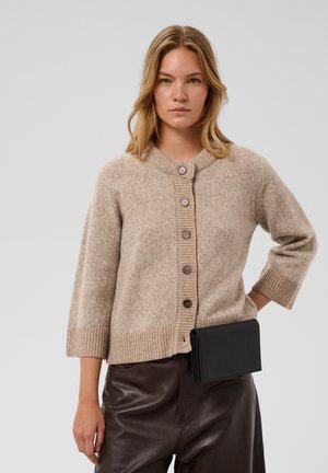 Femme portant un cardigan beige boutonné avec des manches trois-quarts et un pantalon en cuir marron foncé tenant une pochette noire.