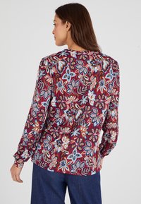 Blusa a maniche lunghe di colore bordeaux con un motivo floreale in blu e arancione. Presenta una linea dritta e un dettaglio arricciato sul collo sul retro.