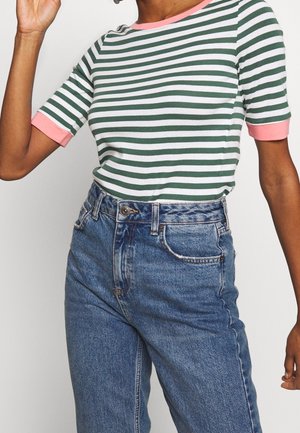 Kvinde iført grøn- og hvidstribet kortærmet skjorte med pink kant og højtaljede blå jeans, poserer med den ene arm hævet.