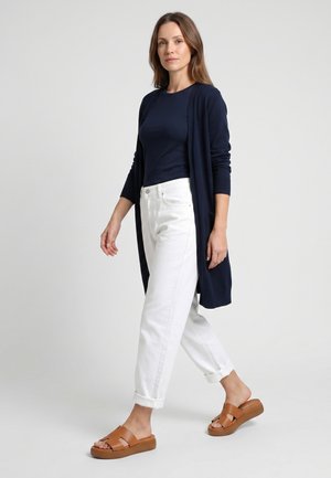 Kvinde går iført en navy cardigan og top, hvide op-rullede bukser og brune åbne slip-on sandaler mod en ensfarvet baggrund.