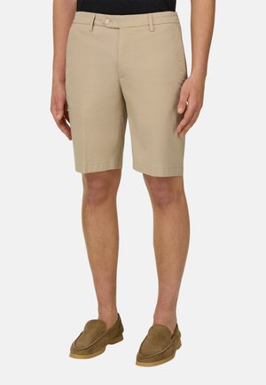 Beige knielange Shorts mit Gürtelschlaufen, getragen von einer stehenden Person, kombiniert mit braunen Wildleder-Loafern und einem schwarzen Oberteil.