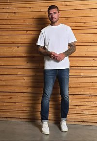 Bílý bavlněný tričko, modré džíny a bílé tenisky. Džíny mají mírné vyblednutí s ohrnutými nohavicemi. Pozadí dřevěné panely.