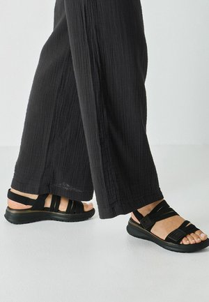 Schwarze Sandale mit Riemendesign, Gummisohle und verstellbaren Verschlüssen. Kombiniert mit lockeren, schwarzen, strukturierten Hosen.