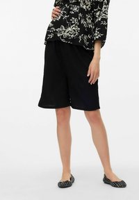 Pantalones cortos negros de corte holgado, fabricados en tela ligera, con un dobladillo recto. Combinados con unos zapatos planos negros con un pequeño detalle de lazo.