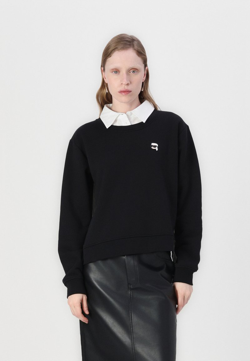 Svart sweatshirt med rund halsringning och ribbade muddar, med en liten logotyp på bröstet. Kombineras med en vit skjorta med krage under.