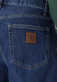 Modré džíny s kapsou na zadní straně, která má viditelné dvojité oranžové švy a hnědou koženou cedulku značky Carhartt.