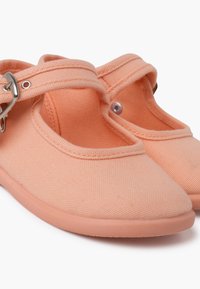Zapatillas de lona melocotón con punta redonda, una correa en el empeine y una suela de goma flexible; parte superior texturizada con detalles de costura.