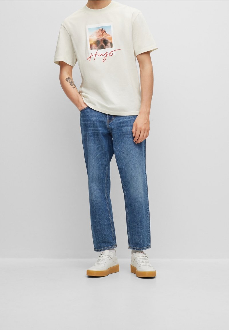 Camiseta crema con un gráfico de montaña y la palabra "Hugs" en rojo, combinada con unos vaqueros rectos azul y zapatillas blancas con suelas marrones.