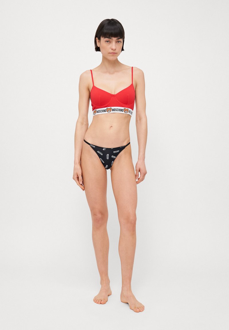 Sarkans bralete ar baltu Moschino logotipa joslu, apvienota ar melniem tangām, kurās atkārtoti redzams Moschino teksts baltā krāsā. Mīksta auduma, minimālistisks dizains.