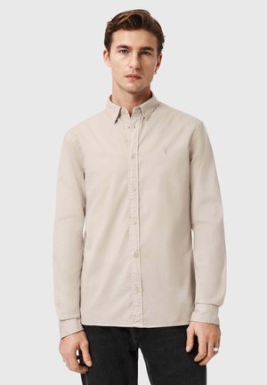 Junger Mann trägt ein beiges langärmeliges Hemd mit Knöpfen und schwarze Hose, steht vor einem schlichten hellgrauen Hintergrund.