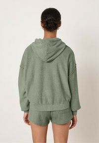 Felpa verde in spugna con cappuccio regolabile, maniche oversize e orlo a costine, abbinata a pantaloni corti coordinati. Dettaglio del tessuto testurizzato visibile.