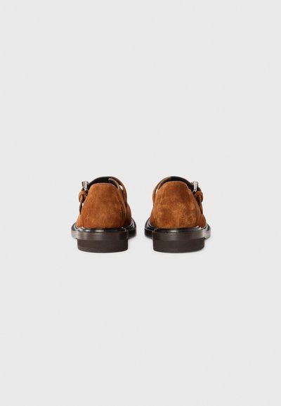 Brown suede shoes with a smooth sole and rounded heel. The back features a decorative strap and subtle stitching details.