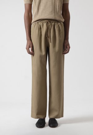 Persona vistiendo pantalones beige de pierna ancha con cintura ajustable y zapatos negros tejidos sin cordones, de pie frente a un fondo claro y sencillo.