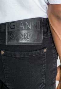 Jeans de denim negro con un parche de cuero texturizado negro etiquetado como "GIANNI KAVANAGH" en la cintura y herrajes dorados en el bolsillo.