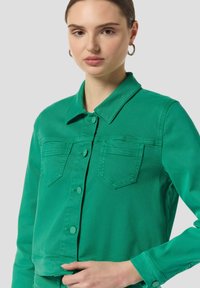 Giacca di denim verde con frontale a bottoni, due tasche sul petto, colletto classico e design leggermente corto con una texture liscia.