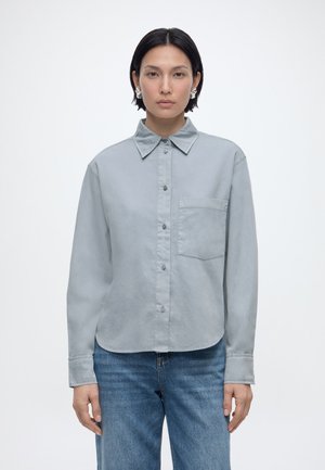 Žena s krátkymi čiernymi vlasmi nosí nadrozmernú svetlosivú košeľu s gombíkmi a vreckom vpredu a modré džínsy, stojaca na bielom pozadí.
