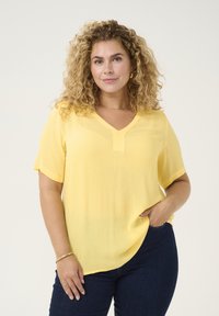 Top à manches courtes, en V, de couleur jaune vif, fabriqué dans un tissu léger et texturé. Présente une coupe décontractée et un subtil détail de couture au niveau du col.
