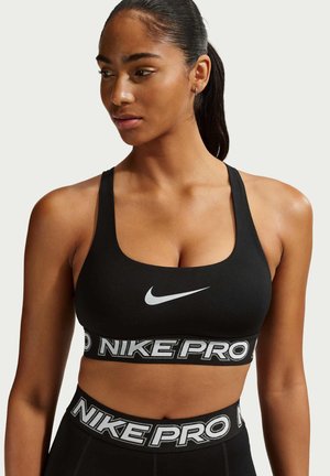 Frau in schwarzem Nike Pro Sport-BH und passenden Leggings mit hoher Taille, die seitlich blickt, vor einem schlichten Hintergrund.