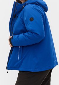 Chaqueta aislante azul con capucha, con cierre de cremallera en la parte frontal, detalles en negro, bolsillos laterales y un emblema de copo de nieve en la manga.