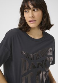 Schwarzes Baumwoll-T-Shirt mit rundem Ausschnitt und kurzen Ärmeln. Auf der Vorderseite steht glänzender Text: "WENN DUZELT, TÄNZE ES HINAUS."