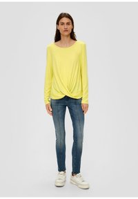 Geel langärmog T-shirt met een geknoopte detail bij de taille, gecombineerd met slim-fit blauwe jeans en witte sneakers.