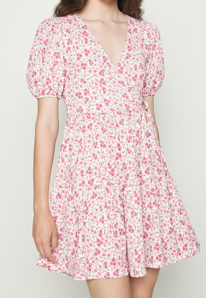 Mujer vistiendo un vestido envolvente blanco con flores pequeñas rosas, mangas abullonadas y una falda acampanada, mostrado desde el cuello hasta la mitad del muslo.