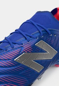 Μπλε ποδοσφαιρικά παπούτσια με υφασμάτινη επένδυση, ζωντανό ροζ πιτσιλιστό μοτίβο, μεταλλικό γκρι λογότυπο και αντίθετο κόκκινο εσωτερικό.