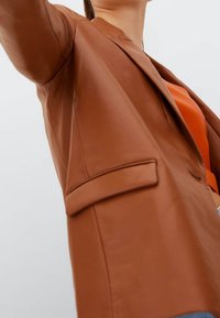 Ruskea nahkapuku, jossa on lovettu kaulus, yksi etutasku ja sileä pinta, puettu oranssin paidan päälle.