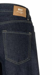 Jean en denim bleu foncé avec une étiquette en cuir beige portant la mention "MAC vintage", des coutures jaunes contrastantes et un design à une seule poche arrière.