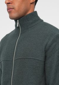 Casaco masculino com fecho éclair na cor verde escuro, feito de tecido texturizado, com colarinho alto, fecho prateado e detalhe de costura horizontal no peito.