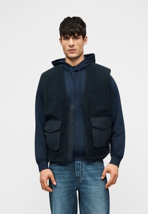 Mann trägt eine marineblaue Kapuzenjacke, darüber eine schwarze ärmellose Fleeceweste mit großen Vordertaschen und blaue Jeans.