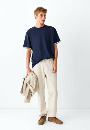 Junger Mann trägt ein marineblaues T-Shirt, beige Hose und braune Schuhe, hält eine beige Jacke und steht vor einem schlichten weißen Hintergrund.