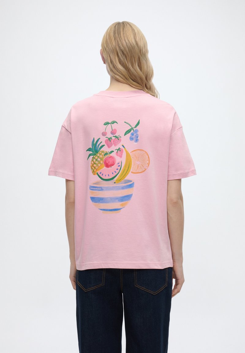 Persona con cabello largo y rubio que lleva una camiseta rosa con una ilustración colorida de un tazón de frutas en la espalda, combinada con jeans oscuros.