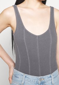 Šedý řasený body s hlubokým výstřihem a tenkými ramínky, v kombinaci se světle modrými džíny, které ukazují detaily šití.