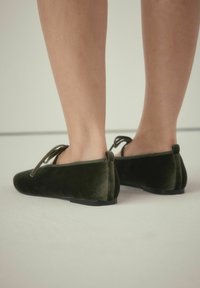 Grønne flade danse sko i fløjl med rund tå, tynde snører og en lille hæl. Skoen har en blød tekstur og en slank silhuet.