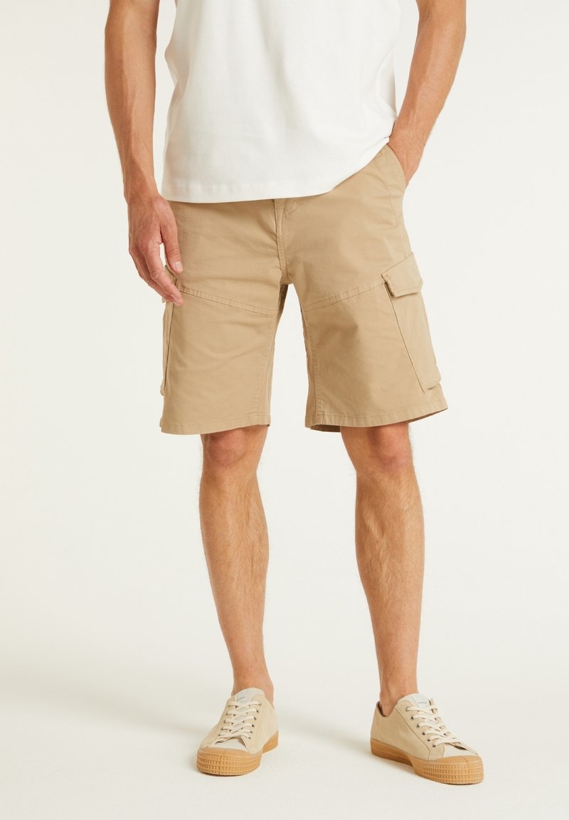 Shorts cargo khaki à coupe droite, avec plusieurs poches latérales et une texture en coton lisse, complétés par des baskets beiges à lacets.