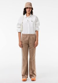 Woman wearing beige patterned bucket hat and matching pants, white buttoned shirt, white shoulder bag, and orange sneakers standing straight.